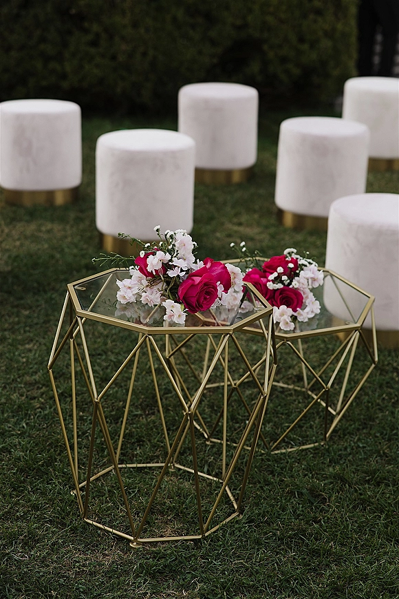 Ceremony seating with white velvet ottomans and a gold geometric glass side table topped with pink roses on a grass lawn by hedges