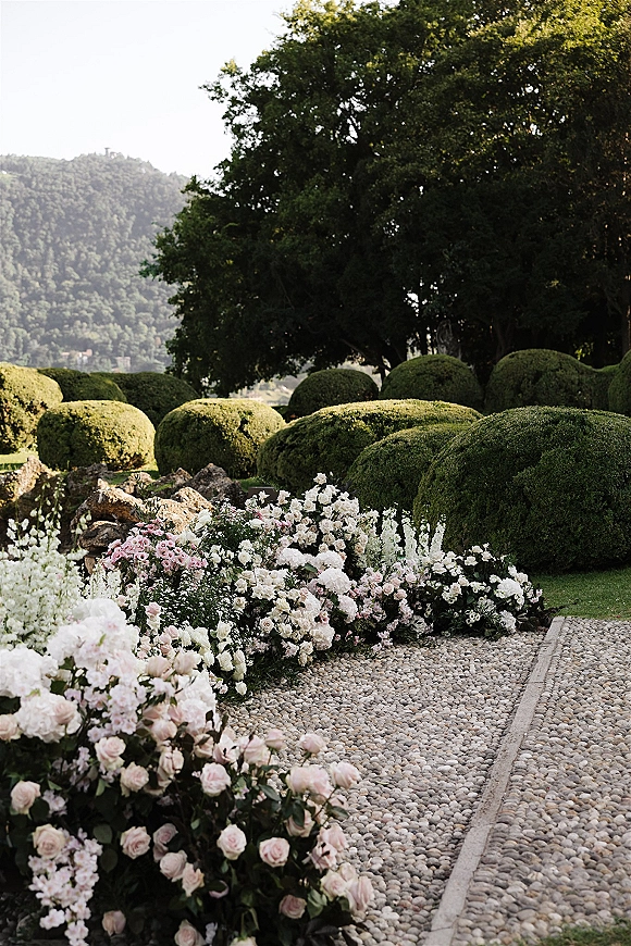 Ceremony aisle decor with wedding aisle flowers in low blush and white rose arrangements on a pebble walkway beside hedges and trees