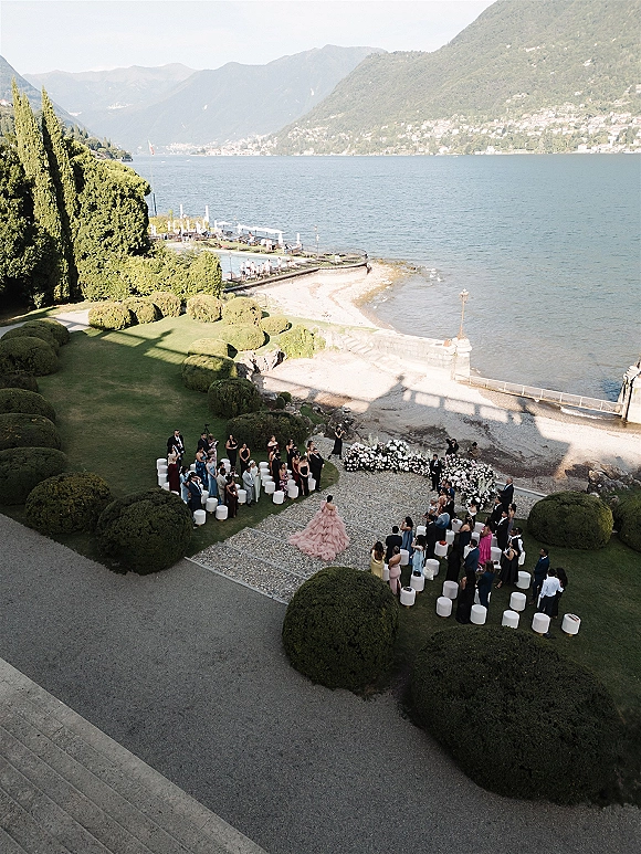 Outdoor wedding ceremony with a floral arch and white aisle chairs on a lakeside lawn, mountains rising beyond the shore
