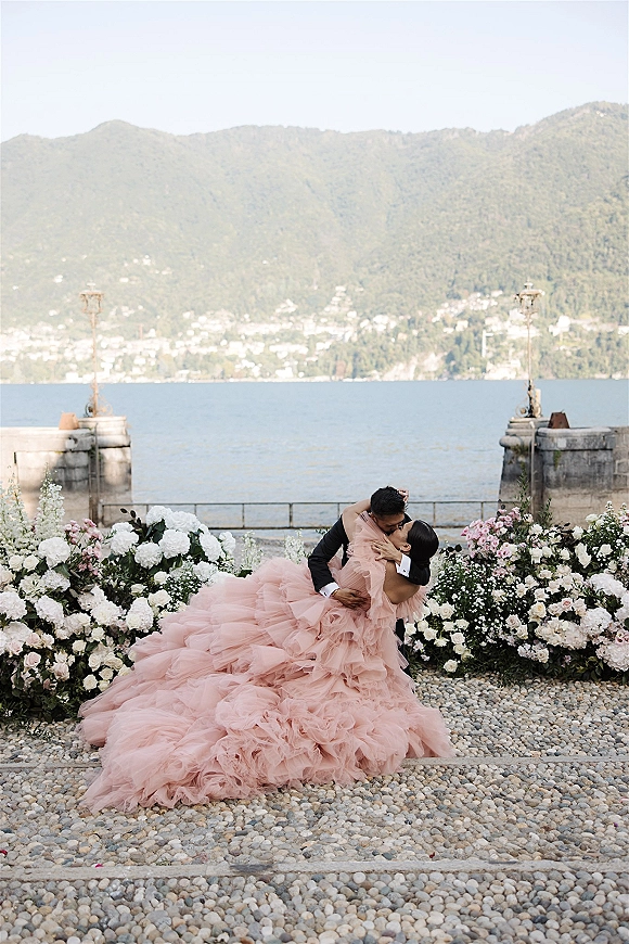Wedding kiss portrait of groom dipping the bride in a blush tulle gown on a stone terrace by a lake with mountain backdrop
