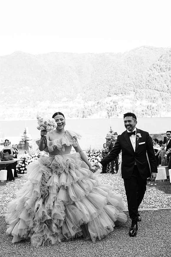 Recessional moment as bride and groom walk the aisle holding hands, bouquet in hand, petals falling with lake and mountains behind