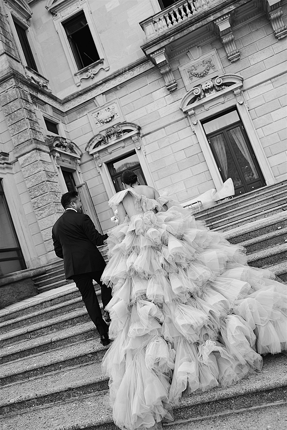 Wedding couple portrait of bride and groom walking up stone steps, her ruffled tulle dress with long train trailing behind, ornate facade ahead