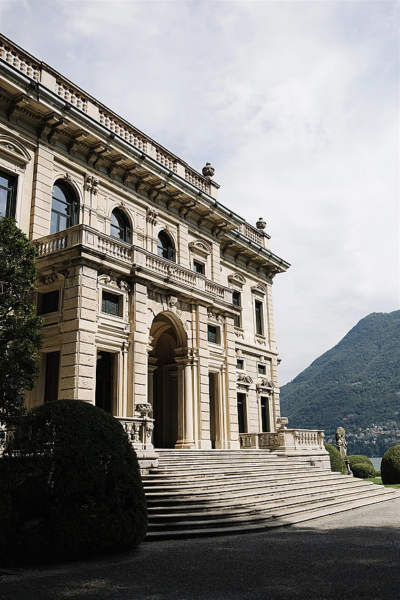 Wedding venue exterior with a grand stone staircase and arched entryway, overlooking a lake and mountains beyond garden shrubs and trees