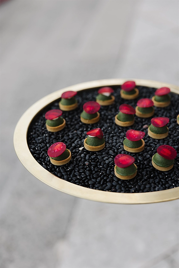 Wedding canapés on a gold serving tray, bite-sized tartlets with green filling and red garnish set over black lentils on gray surface