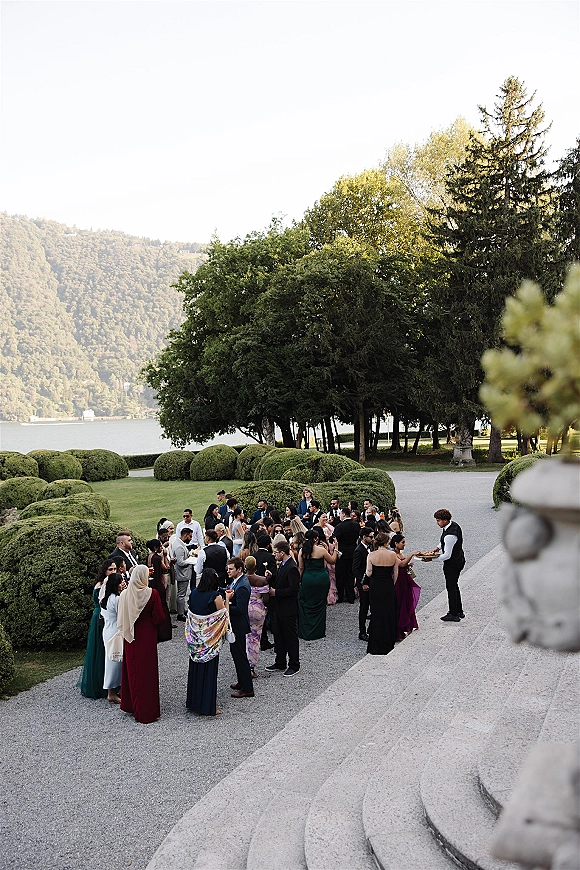 Wedding cocktail hour with wedding guests mingling in formal attire, holding drinks as a server carries a tray on stone steps by a lakeside garden lawn