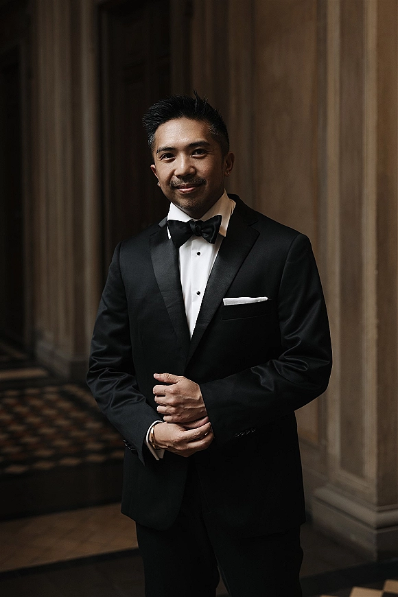 Groom portrait in a black tuxedo with bow tie, hands clasped, standing in a wood-paneled hallway with columns and patterned carpet