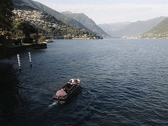 Wedding boat exit with bride and groom on boat, groom driving wooden speedboat as her dress train trails on a mountain lake near village