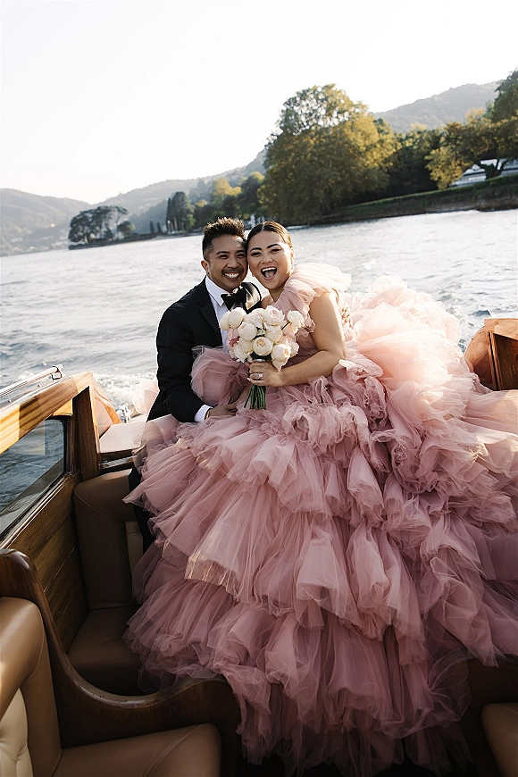 Couple portrait of newlyweds in a wooden boat, bride in blush tulle ball gown holding bouquet, mountains and lake behind