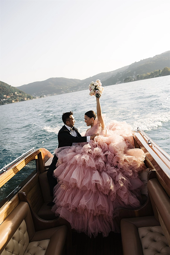 Couple portrait on a classic wooden boat, bride in pink tulle gown lifting bouquet overhead as groom in tuxedo sits on a lake with mountains