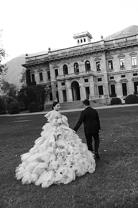 Couple portrait of bride and groom holding hands as she looks back, tulle ruffle gown and veil flowing before a stone mansion doorway