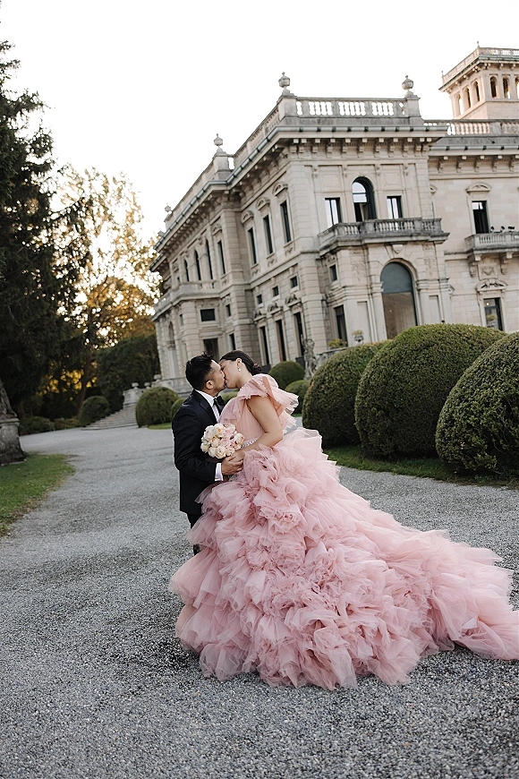 Wedding kiss portrait of bride and groom kissing on a grand estate driveway, her blush pink ruffle ball gown with long train and bouquet