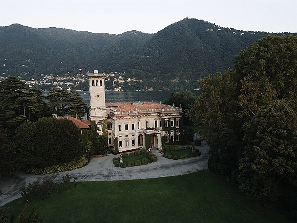 Wedding venue exterior of a historic villa wedding venue with clock tower, ivy walls, terracotta roof, and hedges by a lake and mountains