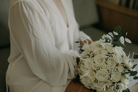 Bridal bouquet of white roses and baby's breath with eucalyptus greenery, tied with ribbon, held in a bridal robe on a sofa indoors