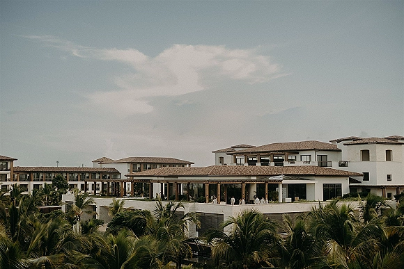 Wedding venue exterior with white stucco buildings and terracotta tile roofs, balconies and pergola, framed by palm trees under cloudy sky