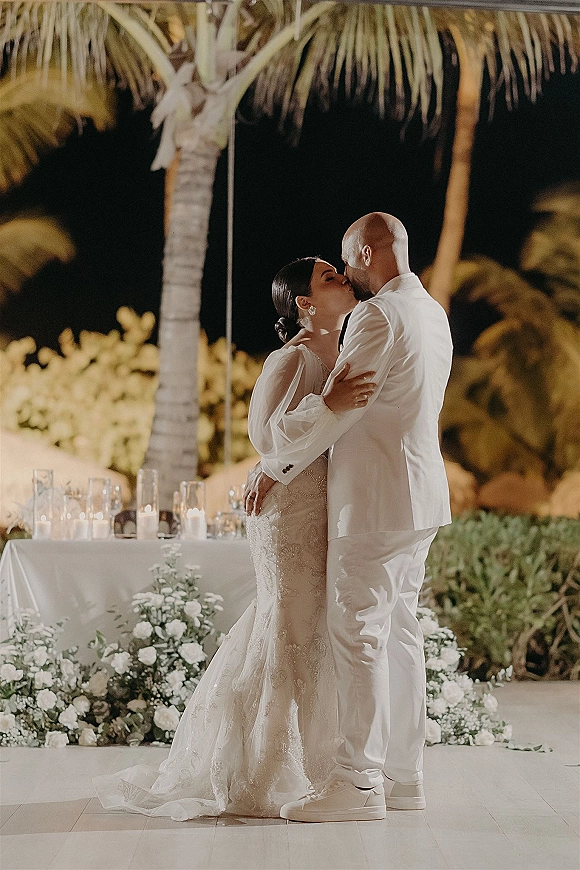 Wedding kiss at a candlelit sweetheart table with white florals and greenery garland, bride in mermaid dress, palm trees behind at night
