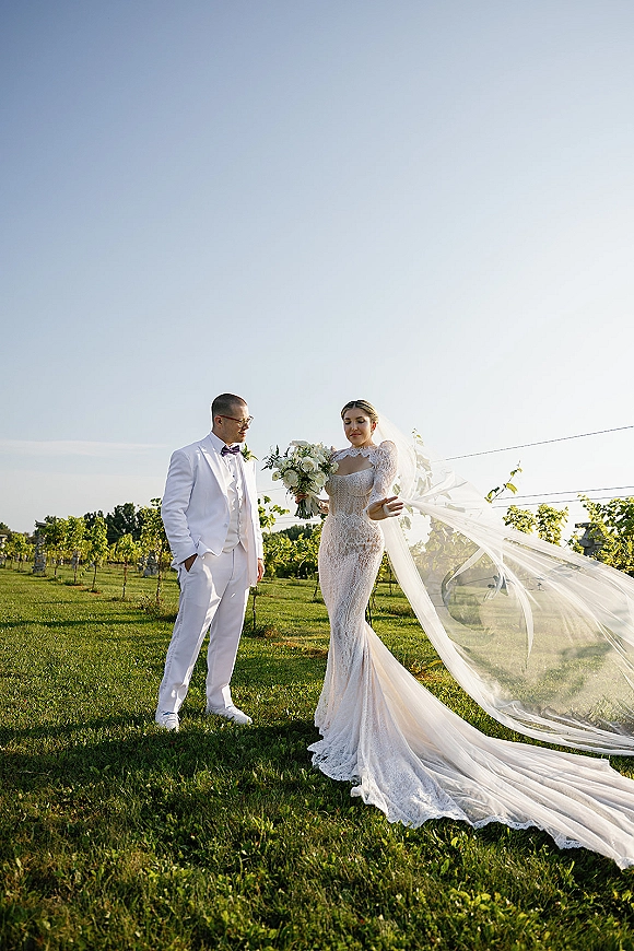 Couple portrait of bride and groom outdoors, her long veil blowing as she holds a white bouquet in vineyard rows under blue sky