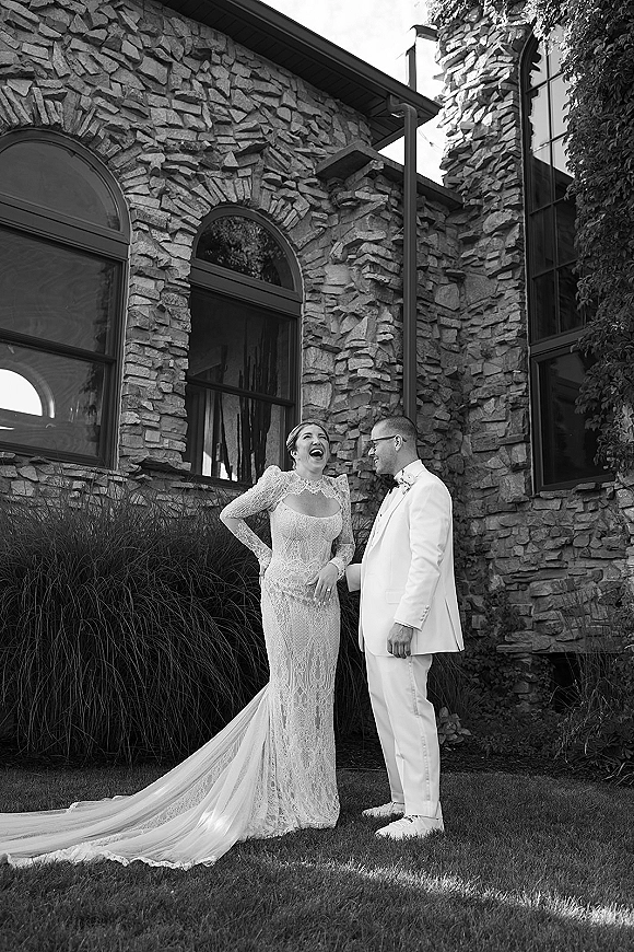 Couple portrait in black and white of bride laughing in a long-sleeve lace gown as groom in white tuxedo looks at her by stone arches