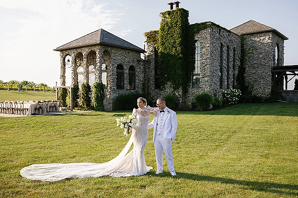 Couple portrait of bride with bouquet and groom in white tuxedo, her lace dress train flowing on lawn by ivy stone villa windows