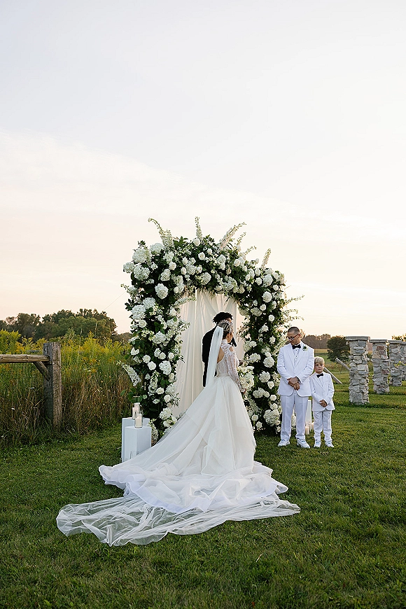 Wedding ceremony with bride and groom under a draped floral arch, veil and long train flowing on a grassy field with lanterns