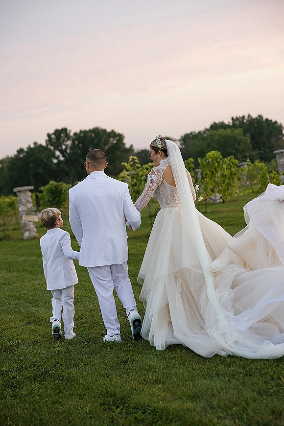 Wedding couple portrait of bride and groom walking away holding hands with a child, her veil and long train trailing in a sunset vineyard