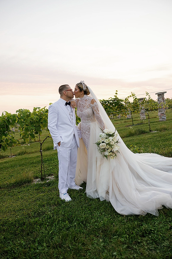 Wedding couple portrait of bride and groom kissing, her lace dress and long veil flowing as she holds white roses in a sunset vineyard