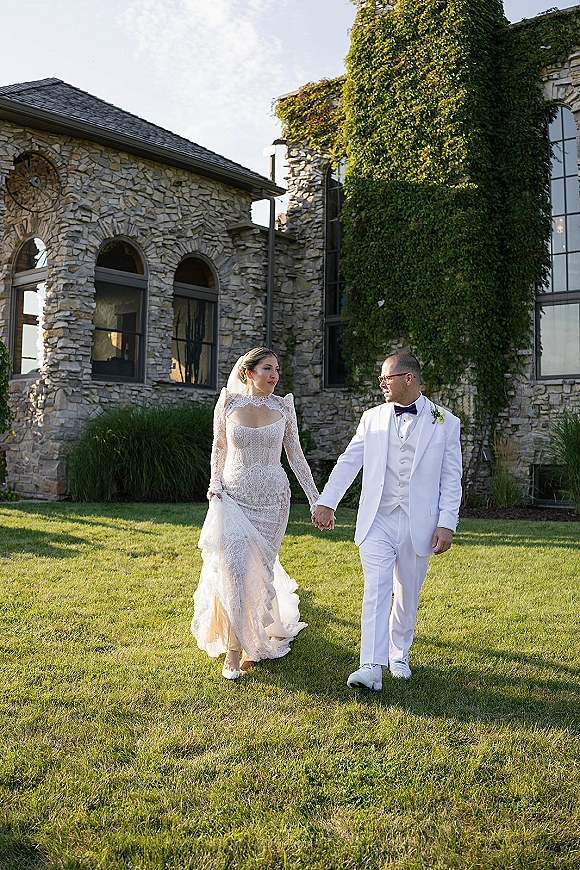 Couple portrait of bride and groom holding hands, bride in lace long sleeve gown and veil, groom in white tuxedo by ivy stone building