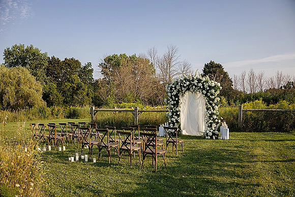 Ceremony setup for an outdoor wedding ceremony with a floral arch and white drapery, wooden chairs, and candle-lined aisle on a lawn