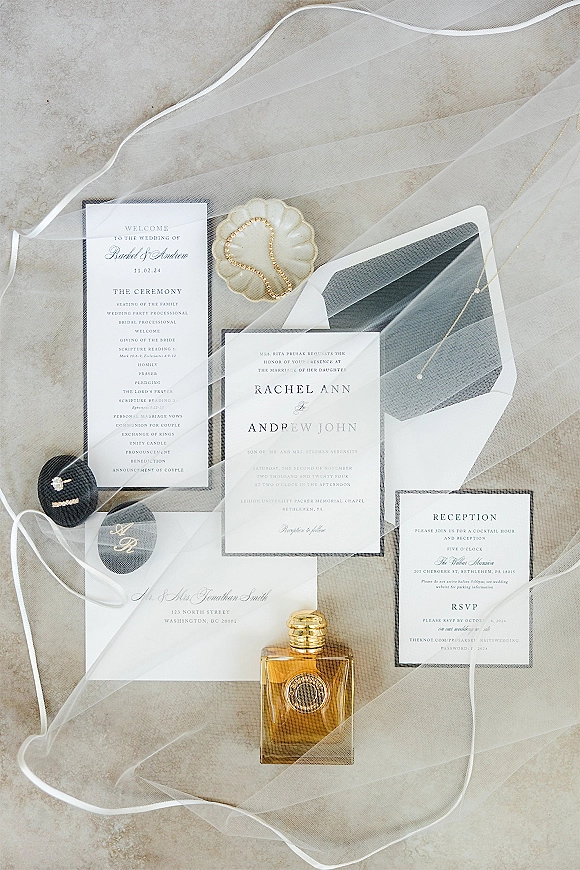 Invitation suite flat lay with black and white wedding invitation cards, envelopes, rings, perfume bottle, and necklace on stone surface