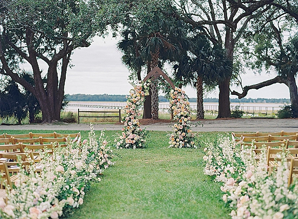 Outdoor ceremony setup with wedding ceremony arch of blush roses and greenery, wooden chairs lining a ribboned aisle by the waterfront dock