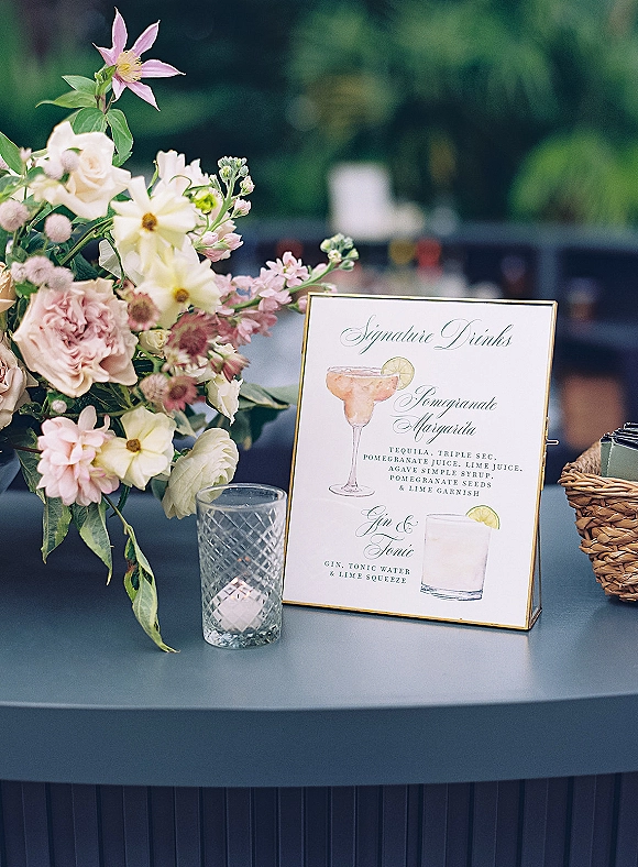 Signature drinks sign in a gold frame with illustrated cocktail menu, beside pastel florals and a candle on a tabletop in greenery outdoors