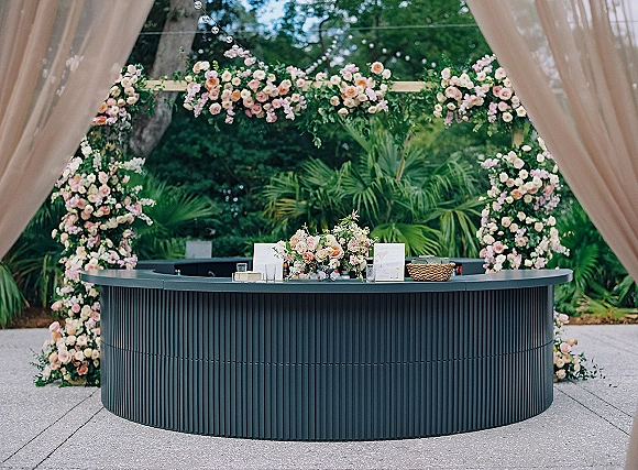 Wedding floral arch with blush rose blooms and greenery, draped fabric, and hanging crystals beside a garden bar on a paved patio