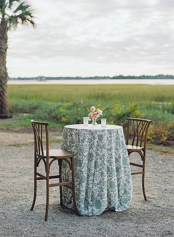 Sweetheart table with a patterned tablecloth, simple floral centerpiece, and glass candle holders by marsh grass and water under a cloudy sky