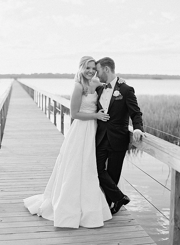 Couple portrait in a black and white wedding portrait, bride in strapless dress embracing groom in tuxedo on a boardwalk by water and marsh grass