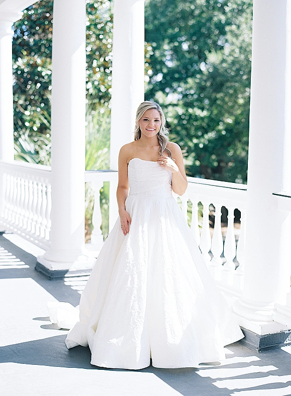 Bridal portrait of a bride in a strapless wedding dress with drop earrings and engagement ring on a sunlit columned porch veranda