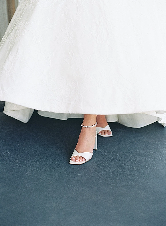 Bridal shoes in white bridal heels with open-toe ankle straps and a rhinestone anklet beside the wedding dress hem on a dark floor