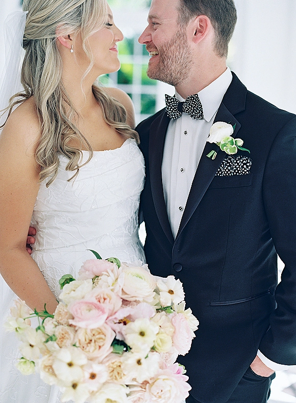 Couple portrait of bride and groom close up, smiling at each other as she holds a bouquet by a bright window with greenery