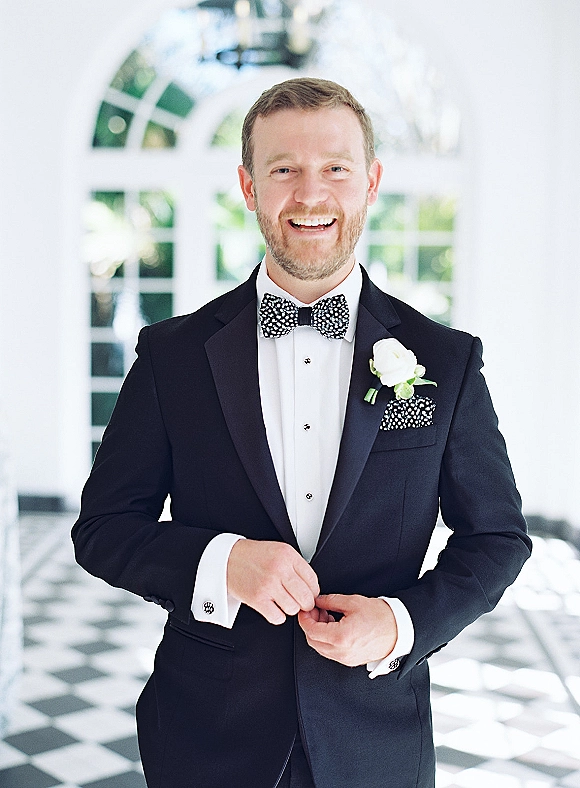 Groom portrait in a black tuxedo with bow tie, boutonniere, and pocket square, standing in a white hallway with arched windows