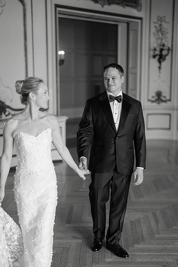 Couple portrait of bride and groom holding hands, walking in a classic ballroom with paneled walls and ornate sconces, strapless lace dress