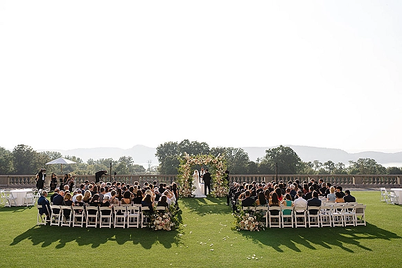 Wedding ceremony with bride and groom at a floral arch, guests in white chairs on a lawn with stone balustrade and hills beyond