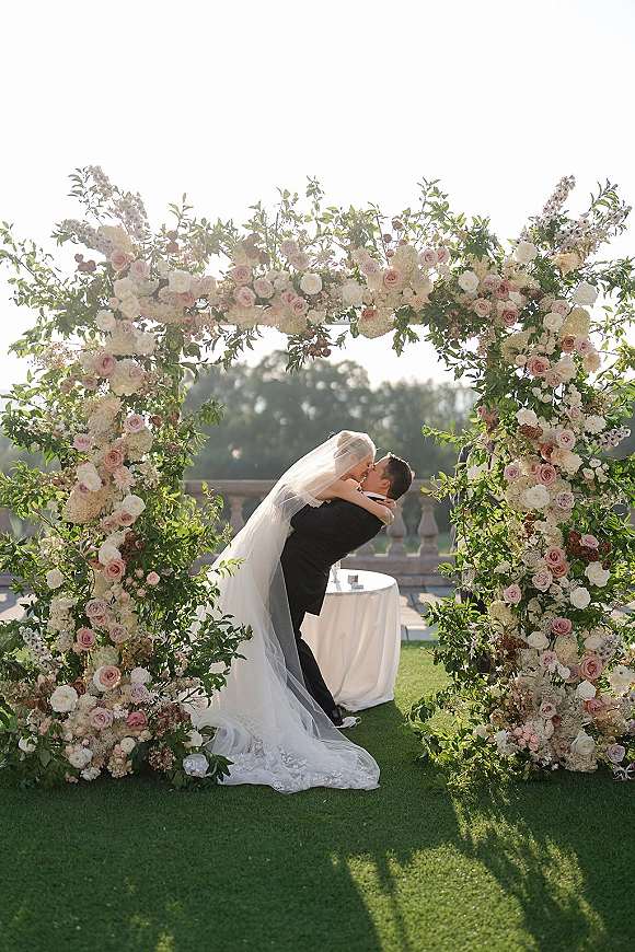 Wedding kiss portrait of bride and groom dipping in embrace beneath a round rose and hydrangea floral arch on a lawn with stone balustrade