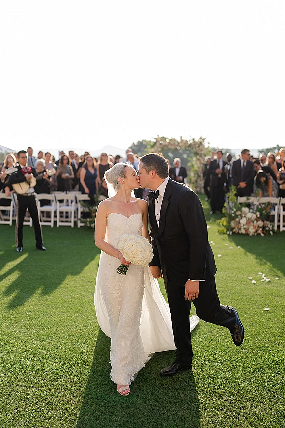 Wedding kiss portrait of bride and groom kissing in a black tuxedo and lace gown, holding a white rose bouquet on an outdoor aisle with guests
