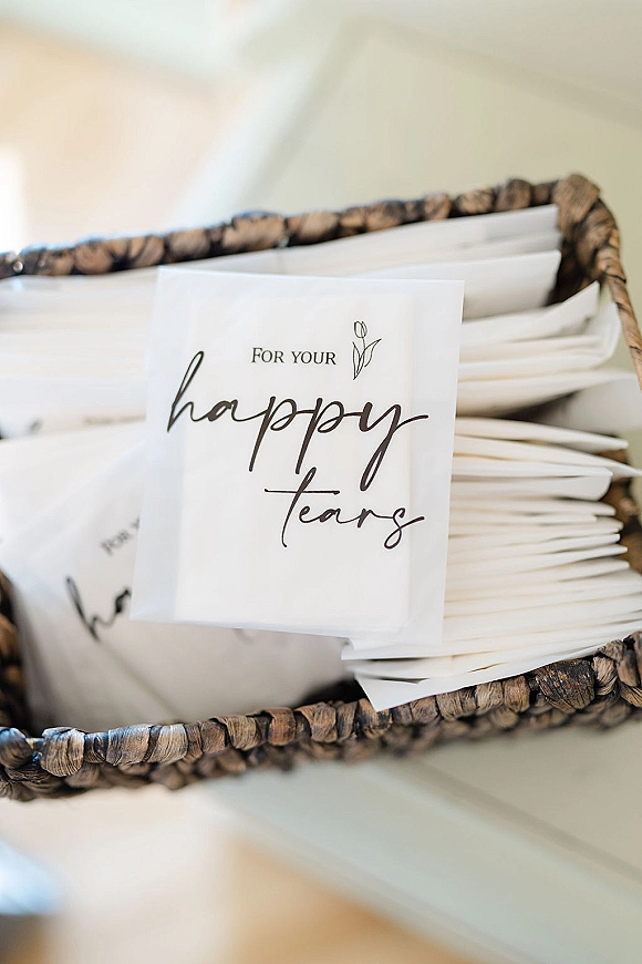 Happy tears bags with calligraphy labels on paper tissue packets, arranged in a woven basket on a table surface