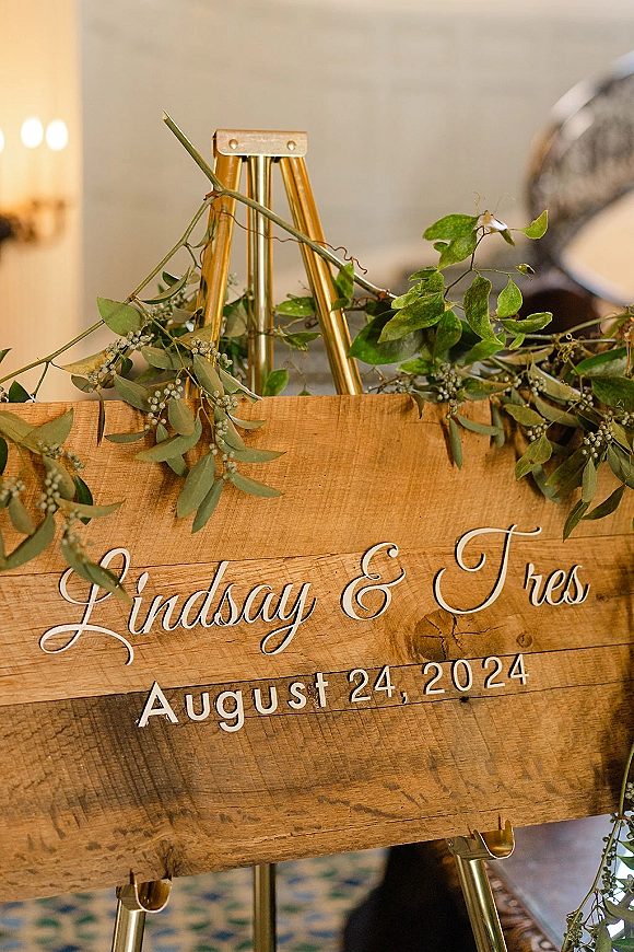 Wedding welcome sign with calligraphy lettering on a wood sign, greenery garland accent, displayed on a gold easel indoors with soft lights