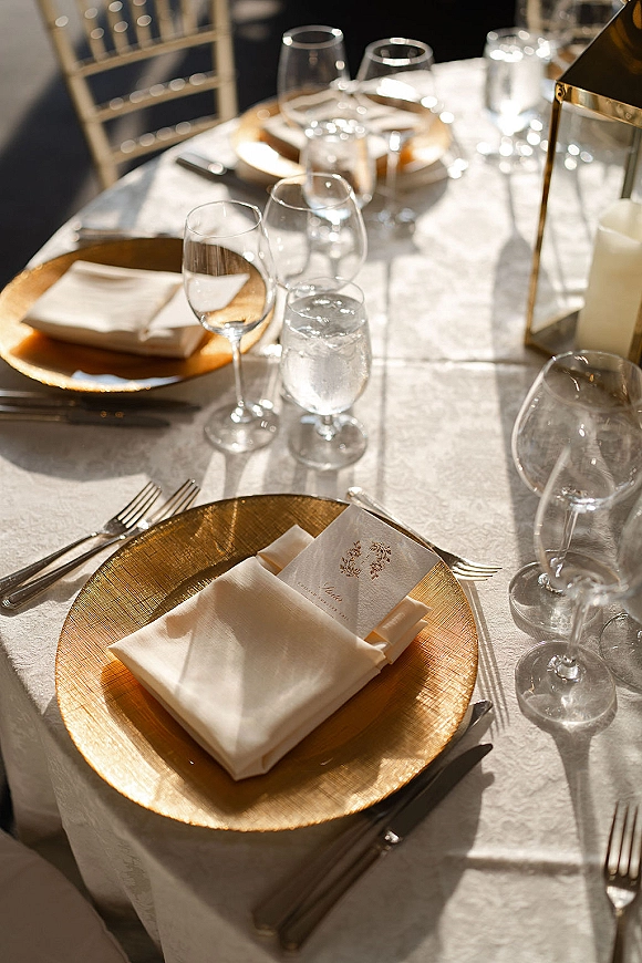 Reception tablescape with gold charger wedding table details, folded napkins and place cards beside wine glasses, sunlit lantern centerpiece