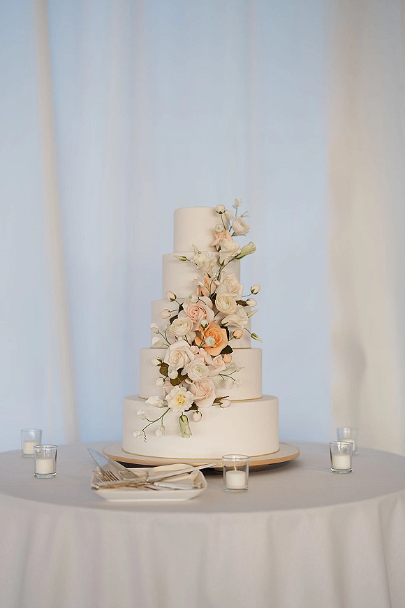 Wedding cake with a white tiered wedding cake design, sugar flower cascade on a stand, surrounded by votive candles and plates before white drapery