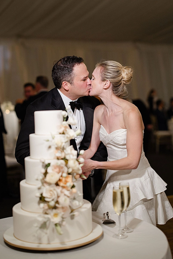 Wedding cake cutting as bride in a strapless gown and groom in tux slice a white tiered cake with blush roses in a draped reception room