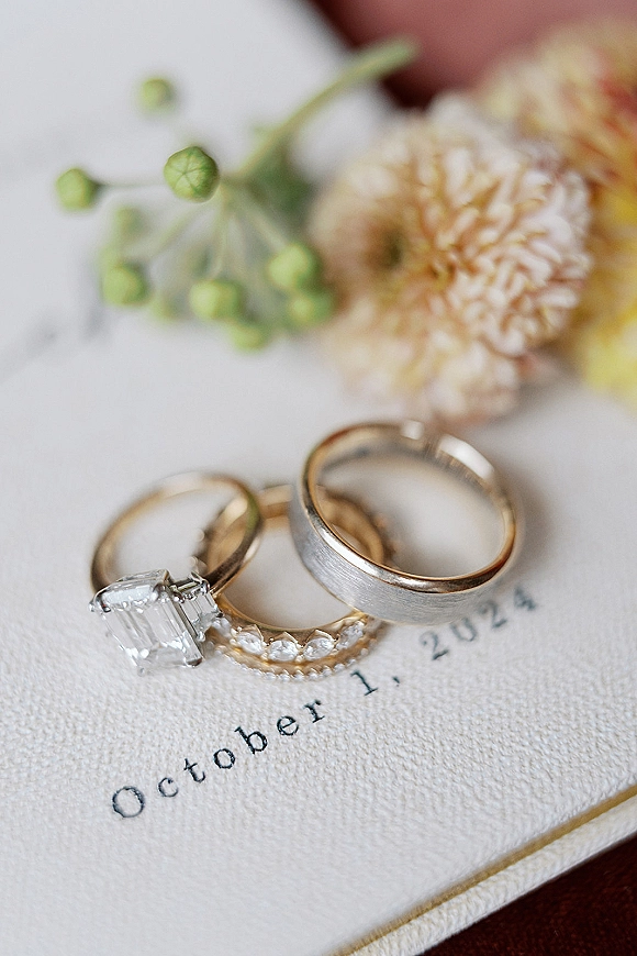 Wedding rings and emerald cut engagement ring close up resting on a wedding invitation with greenery on a wooden surface