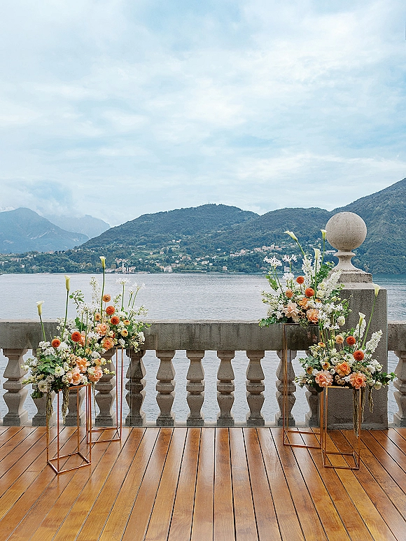 Wedding ceremony decor with rose and calla lily florals on gold pedestal stands, framed by a stone balustrade and lakeside mountains backdrop
