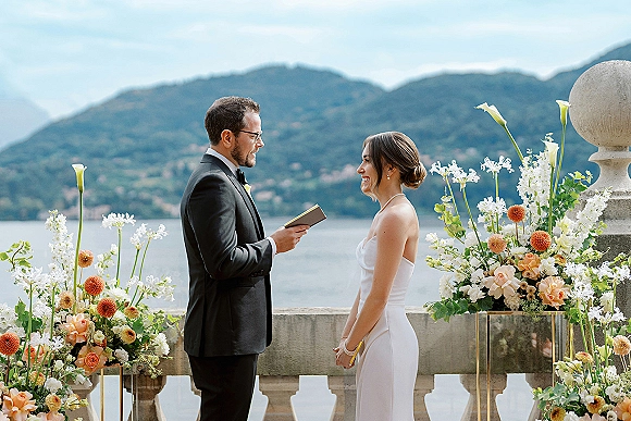 Wedding vows shared as groom reads from a vow book beside bride in strapless satin gown on a stone terrace with lake and mountains beyond