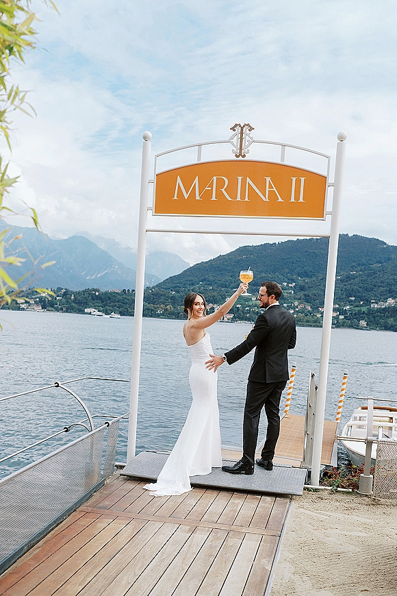 Couple portrait of bride and groom toast with champagne coupe on a dock, mountains and lake behind, marina sign near railing